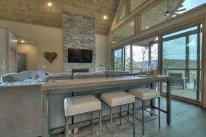 A modern kitchen with a stone fireplace, a large TV, and a bar area with stools. - 4 Dancing Bears|Mtn Views+Hot Tub+Interior Design (Blue Ridge)