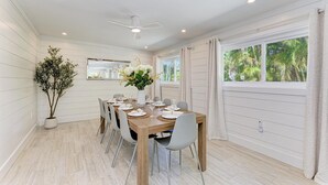 Un comedor con una mesa de madera dispuesta para cuatro, paredes blancas, ventilador de techo y un amplio ventanal con cortinas.