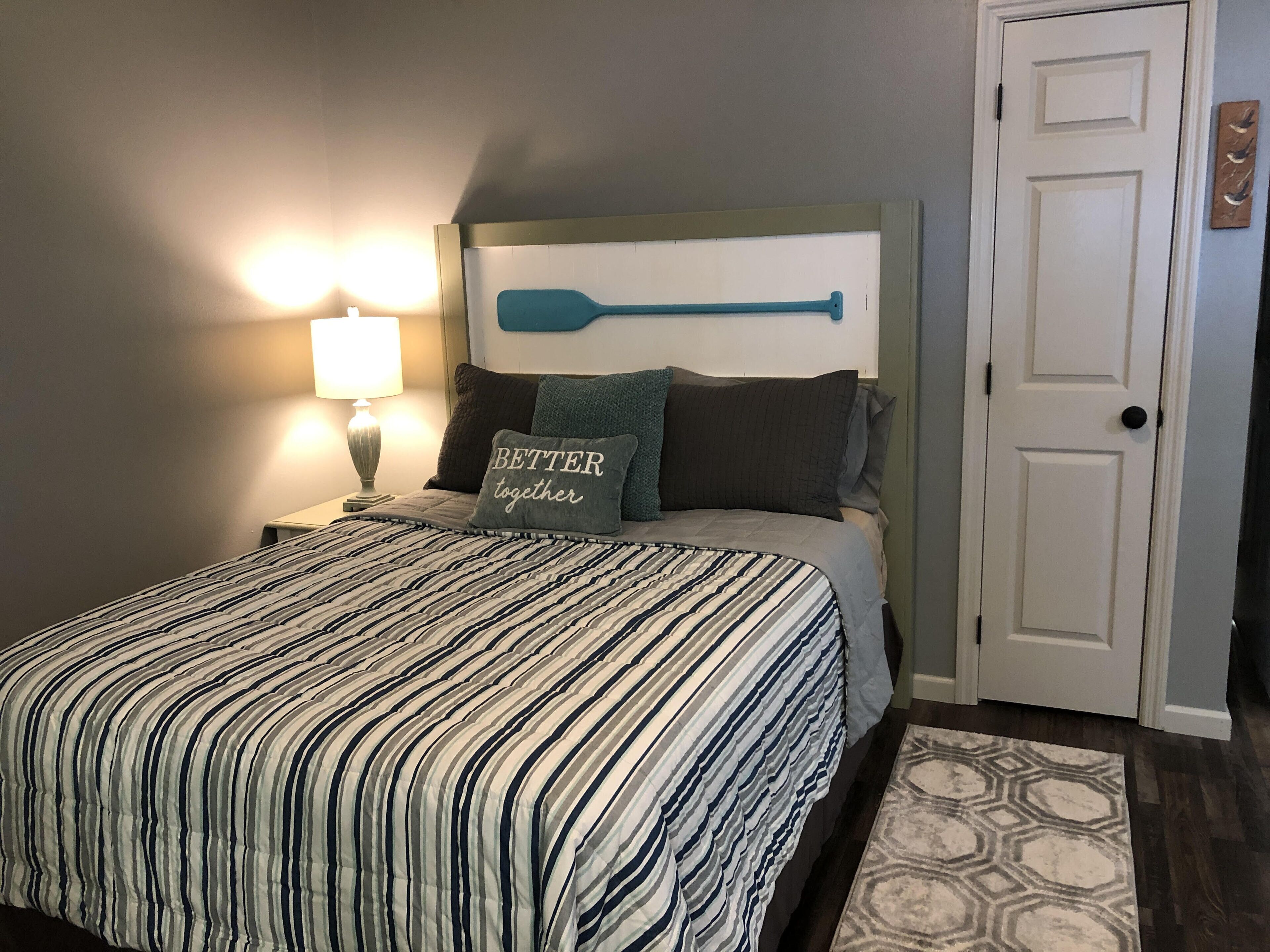 Una cama bien hecha con ropa de cama a rayas, un cabecero fijado a la pared, una lámpara de noche y un almohadón con la inscripción 'BETTER together'.