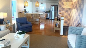 A modern living room with a blue sofa, a white coffee table, and a kitchen area with white cabinets and a stainless steel refrigerator. - 4 bedroom, 2 bath home with 3 porches, & steps from the beach in Atlantic Beach (Atlantic Beach)