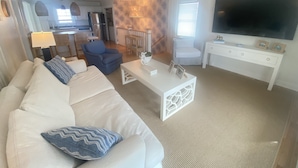 A living room with a white sofa, blue armchair, and a coffee table. There is a TV on the right and a kitchen in the background. - 4 bedroom, 2 bath home with 3 porches, & steps from the beach in Atlantic Beach (Atlantic Beach)