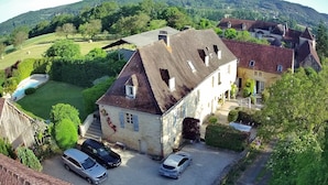 A stone building with a steep roof, a driveway with two cars, and a surrounding green landscape.