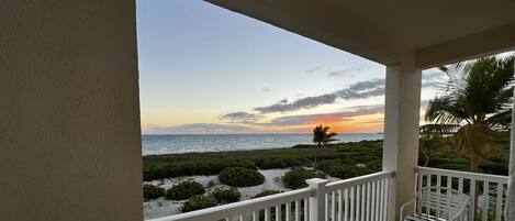 A balcony with a view of the ocean at sunset, white railings, and a chair.