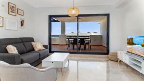 Ein modernes Wohnzimmer mit einem grauen Sofa, einem weißen Couchtisch und einem Fernseher auf einem weißen Schrank. Ein großes Schiebeglasfenster führt zu einem Essbereich im Freien.