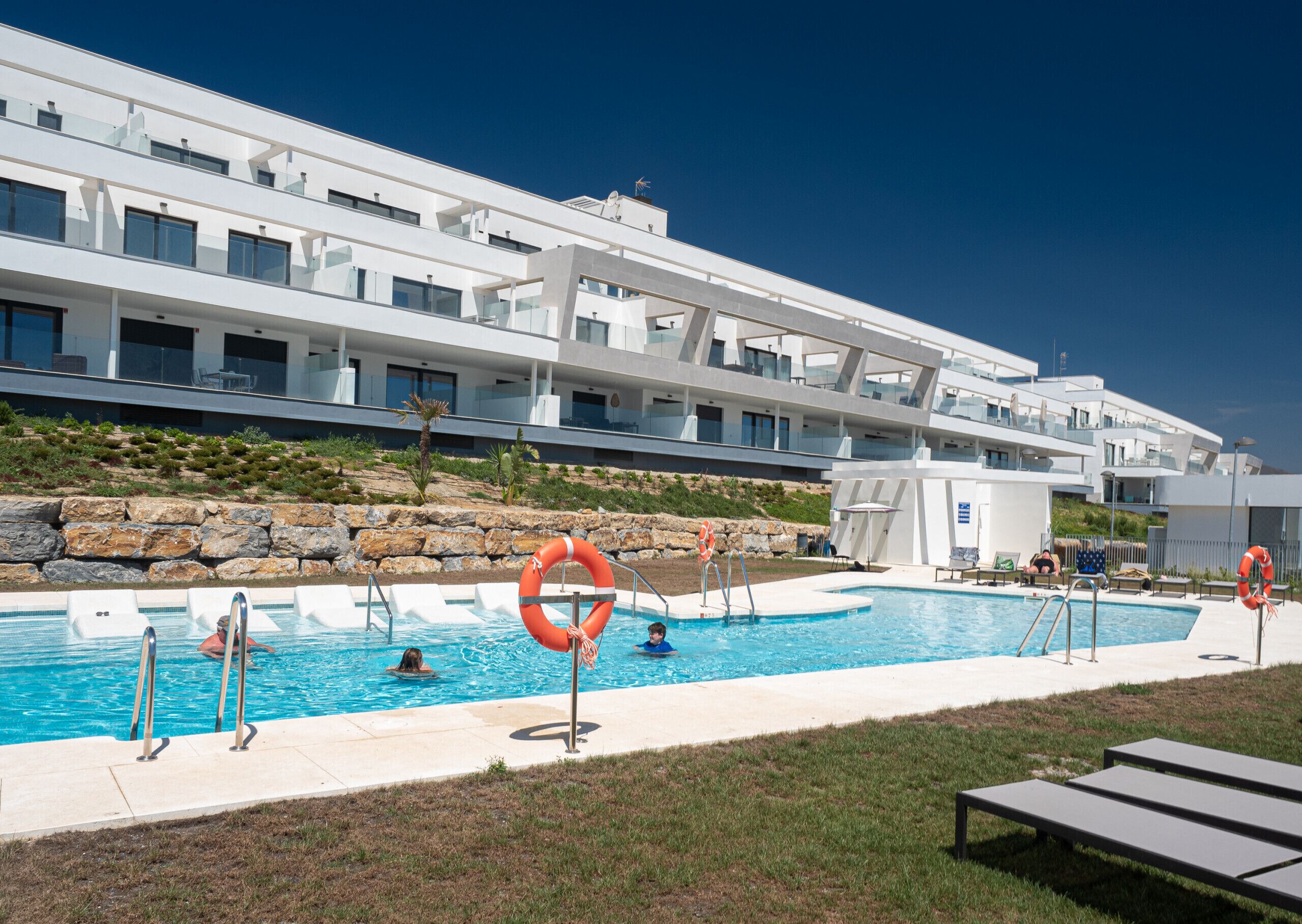 A modern hotel with a swimming pool, lounge chairs, and a rocky hillside backdrop.