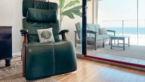 Una silla de cuero verde con un cojín, ubicada en una habitación con piso de madera y vista a la playa a través de un ventanal.