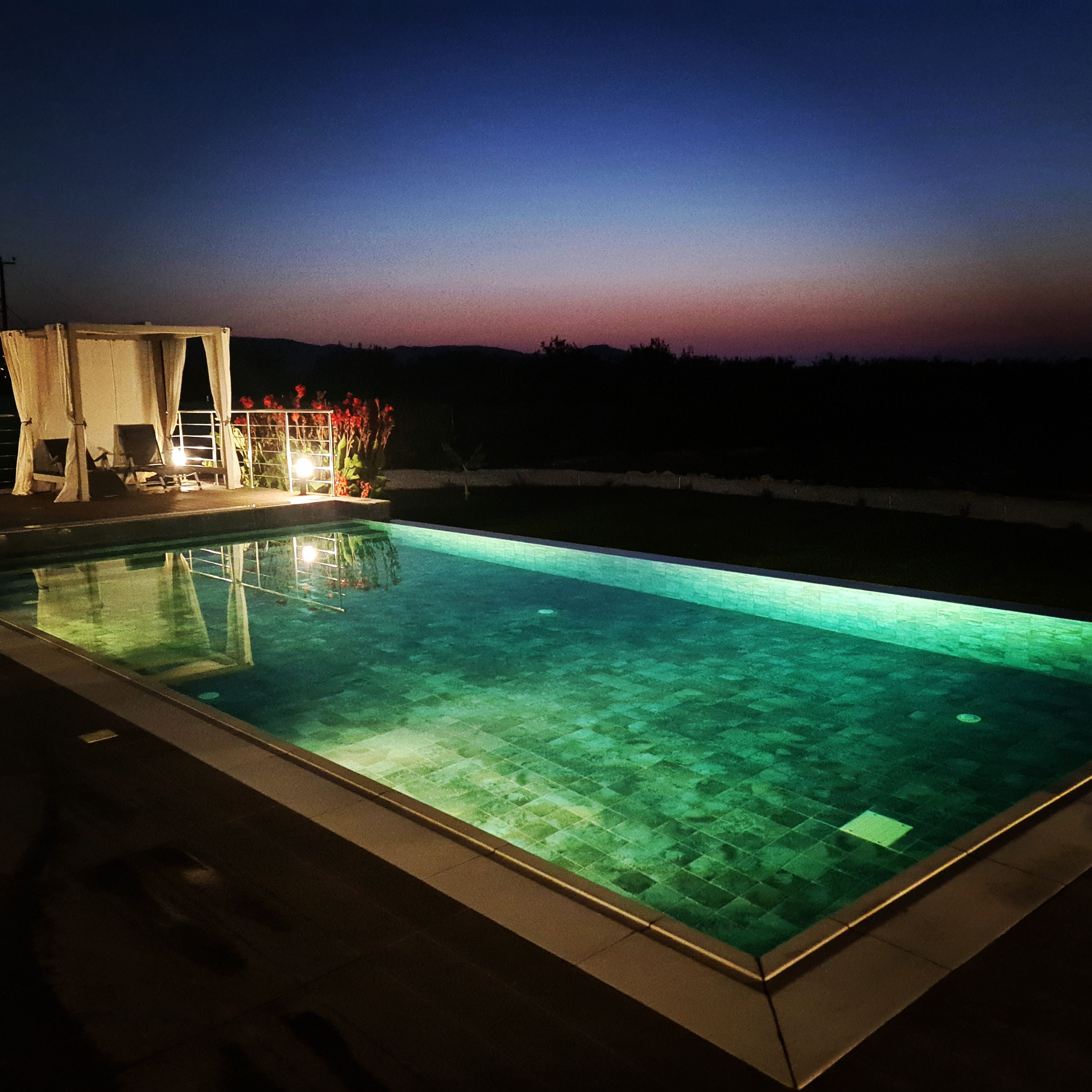 A rectangular swimming pool with clear water, surrounded by a patio area with chairs and a small table, and a covered seating area with curtains.
