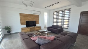 A modern living room with a sectional sofa, a flat-screen TV, a wooden entertainment unit, a grey armchair, and a large window with blinds. - Modern Luxury Villa near Lachi (Prodromi)