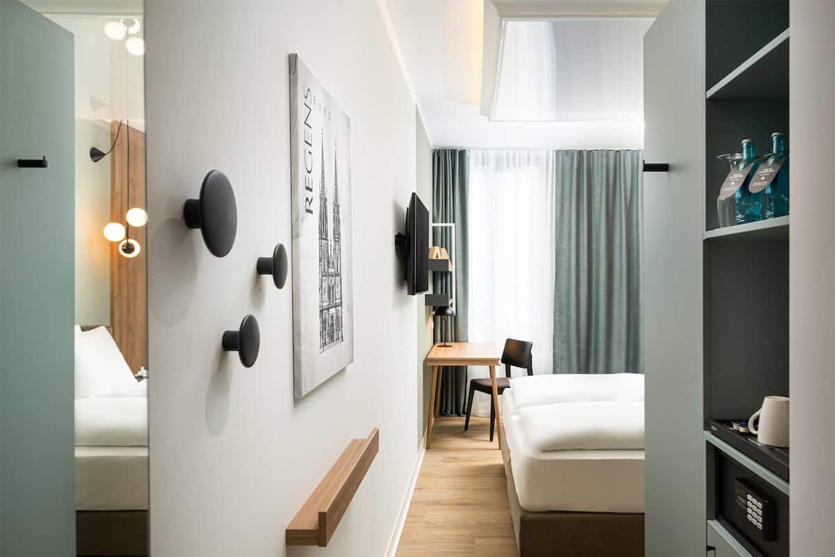 A modern hotel room with a bed, a desk, a chair, a TV, and a shelf with bottles.