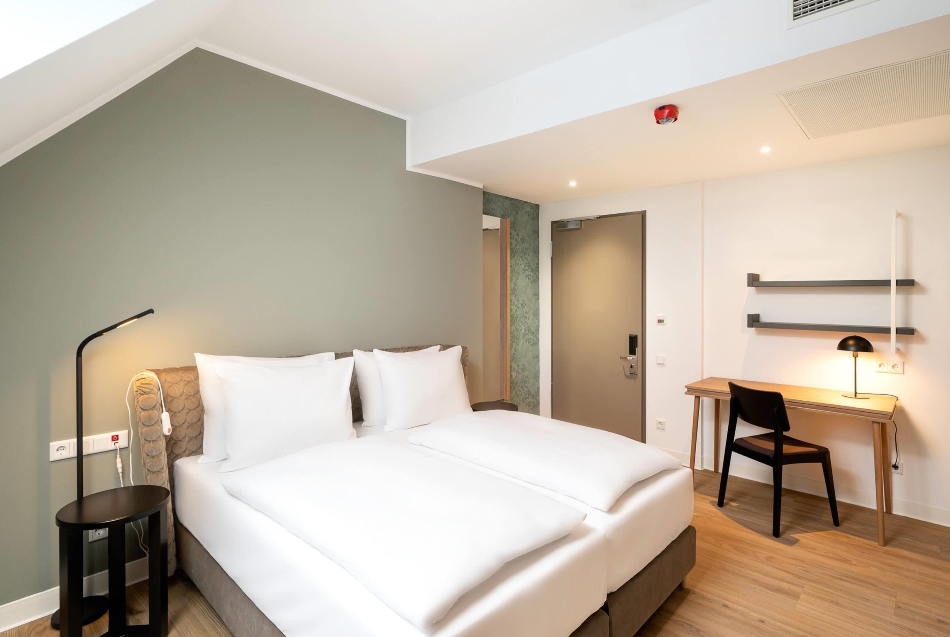 Ein modernes Hotelzimmer mit Bett, Nachttisch, Lampe und Schreibtisch mit Stuhl.