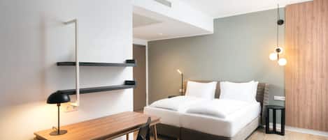 A modern hotel room with a bed, a desk, and a chair. There is a bedside table with a lamp, a wall-mounted shelf, and a hanging light fixture.