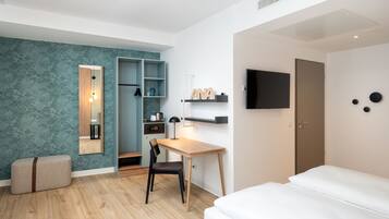A modern hotel room with a wooden desk, a chair, a bed, a wardrobe, a TV, and a shelf with a nameplate.