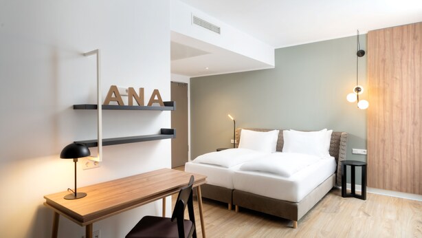 A modern bedroom with a bed, a desk, and a chair. There is a shelf with the letters "ANA" on it, a lamp, and a bedside table with a lamp. - elaya hotel regensburg city center, Trademark by Wyndham (Regensburg)