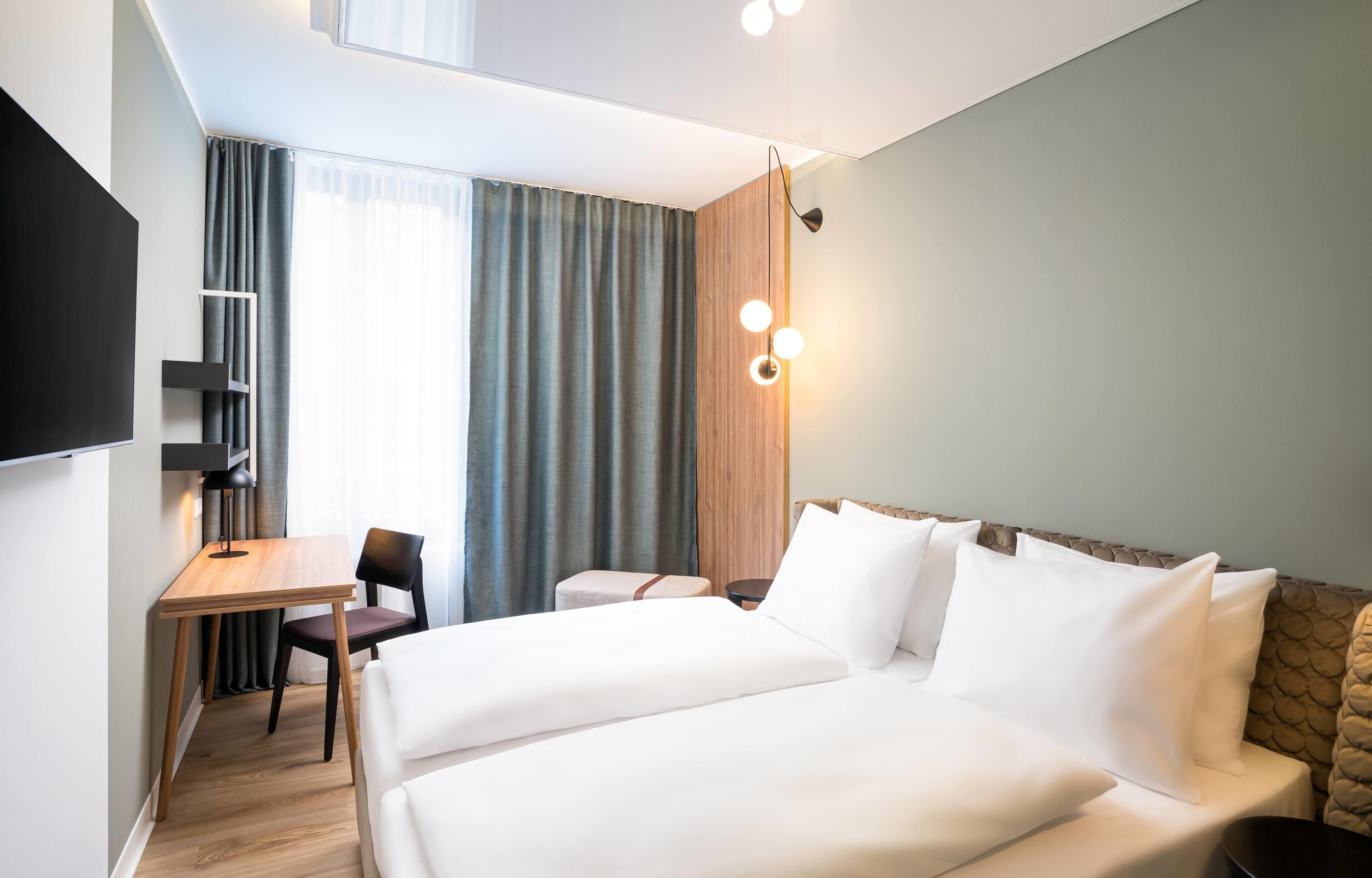 Une chambre d’hôtel avec un lit, un bureau, une chaise, une télévision et une fenêtre avec des rideaux.