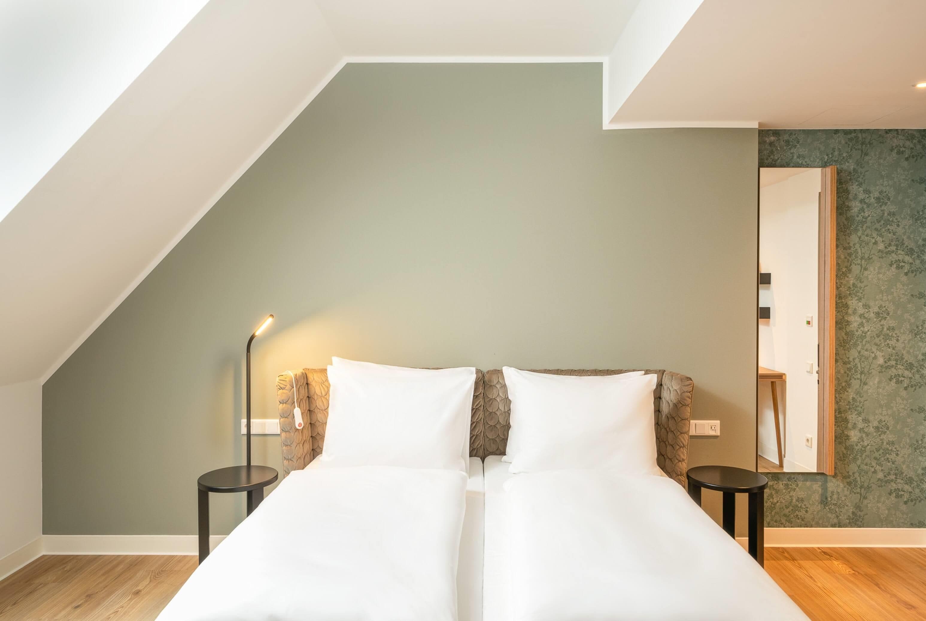 A room with a sloped ceiling, a bed with white bedding, two bedside tables, a floor lamp, and a mirror on the wall.
