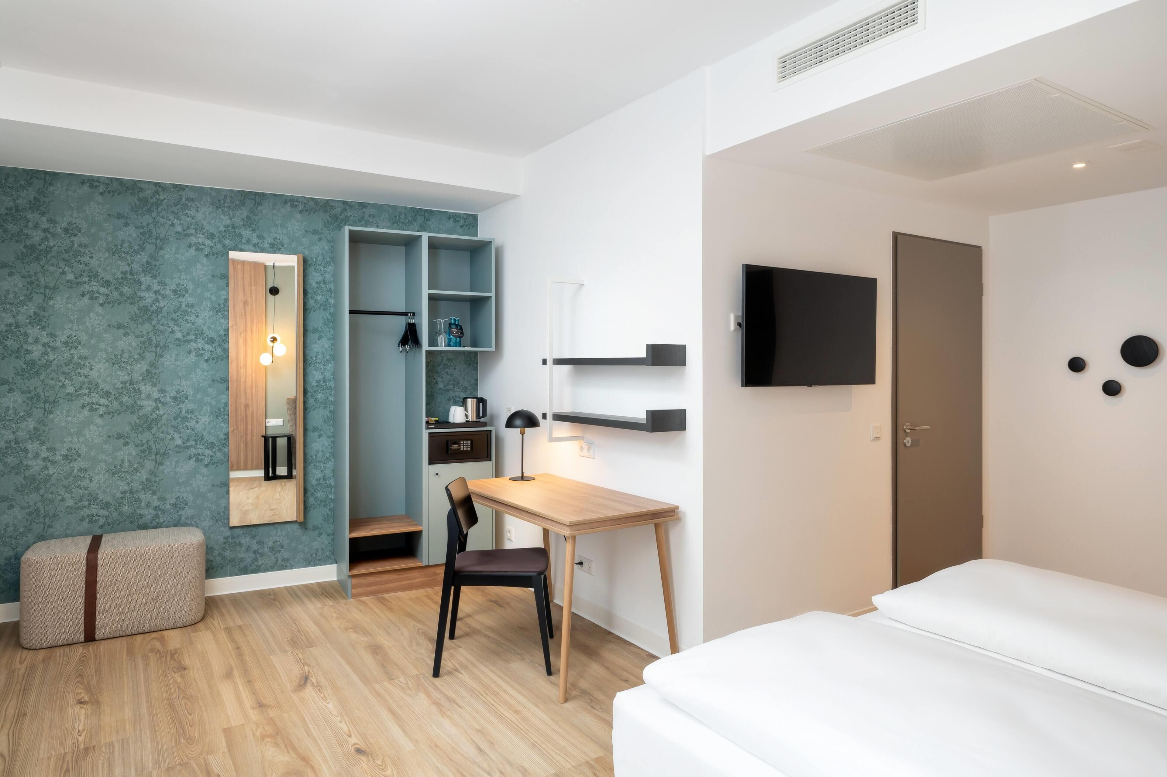 A modern hotel room with a wooden desk, a bed, a TV mounted on the wall, and a wardrobe with built-in shelves.