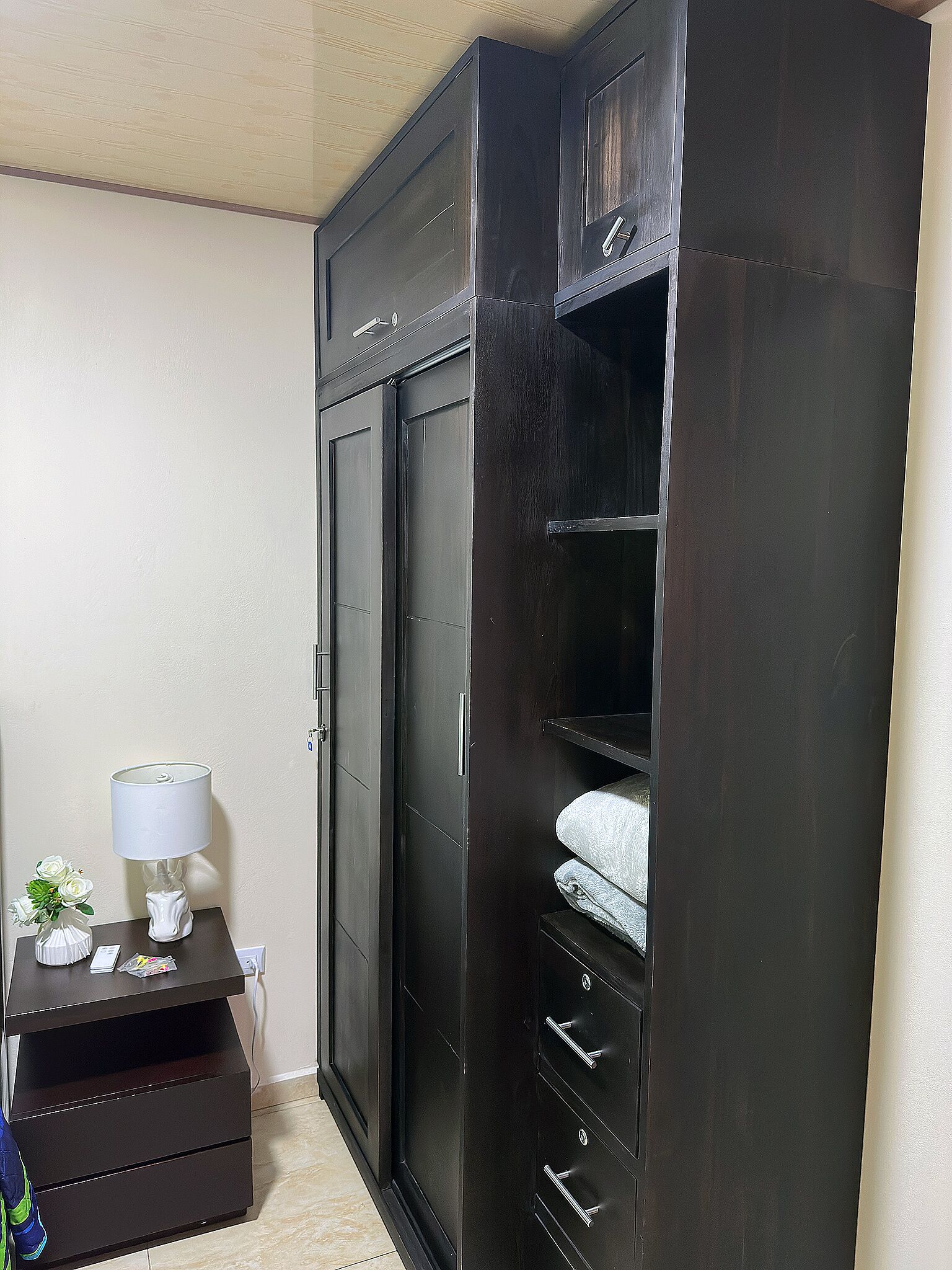 Une grande armoire en bois sombre, dotée de plusieurs portes et de tiroirs, une petite table avec une lampe et un vase de fleurs, et un sol carrelé.