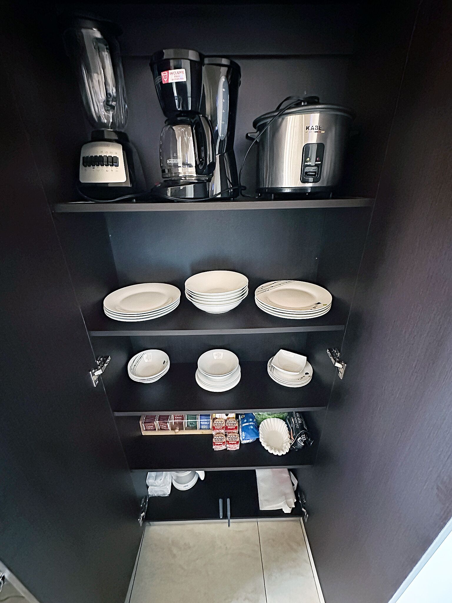A kitchen with a coffee maker, blender, and rice cooker on the upper shelf, and plates and utensils on the lower shelves.