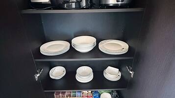A kitchen with a coffee maker, blender, and rice cooker on the upper shelf, and plates and utensils on the lower shelves.