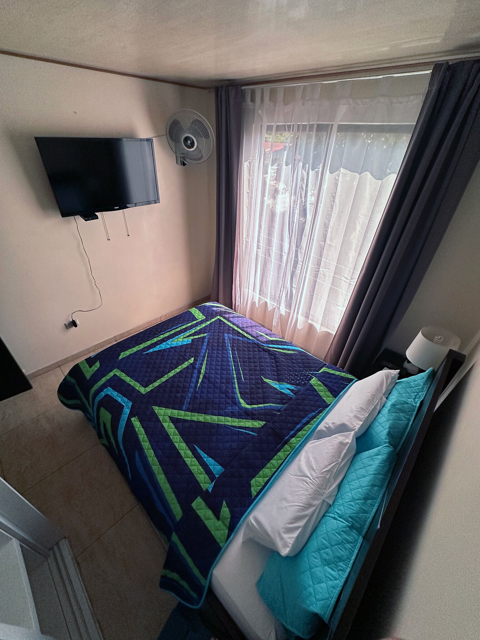 A small, neatly made bed with a patterned bedspread, a wall-mounted TV, and a window with curtains.