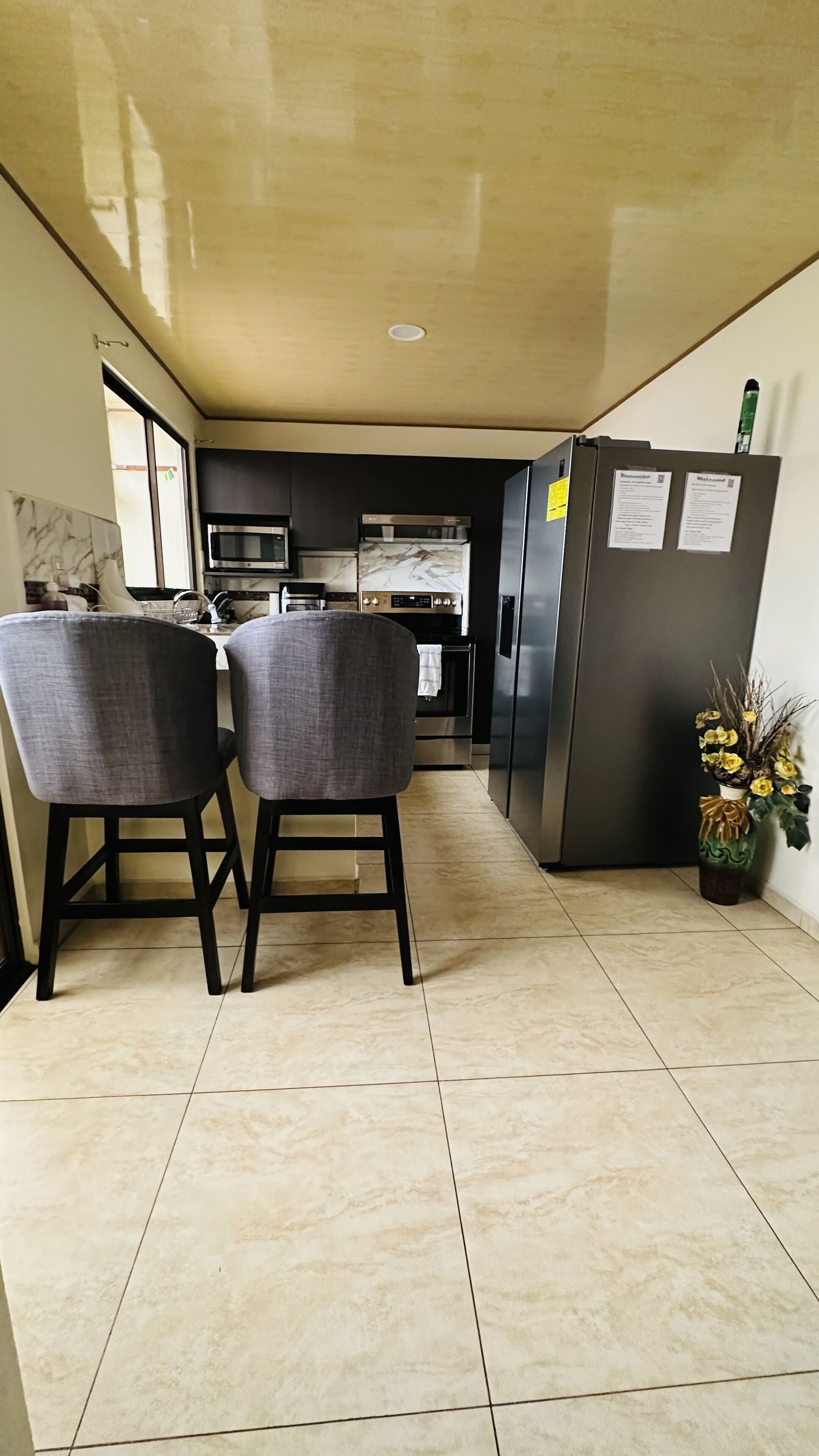 Una cucina moderna con forno a microonde, forno e frigorifero. Sono presenti due sedie grigie e un vaso con fiori gialli.