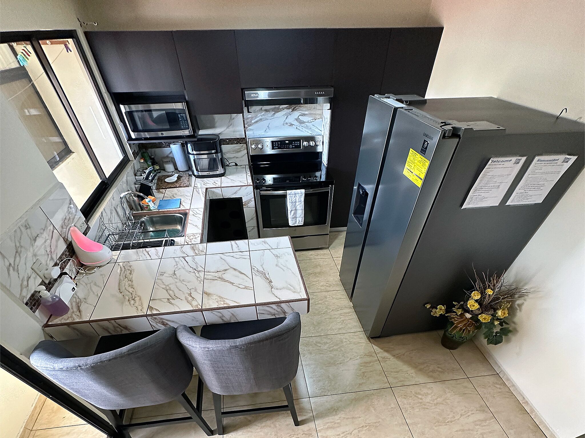 A compact kitchen with a marble countertop, two grey chairs, a stainless steel refrigerator, and a microwave.