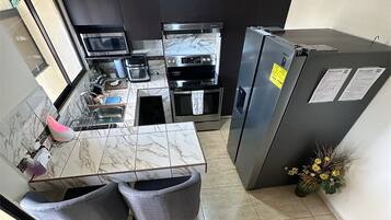 A compact kitchen with a marble countertop, two grey chairs, a stainless steel refrigerator, and a microwave.