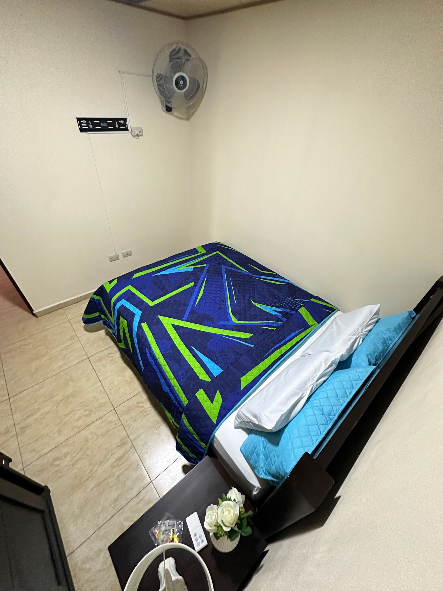 A small, neatly arranged bedroom with a bed, a bedside table, a fan, and a wall-mounted light switch.