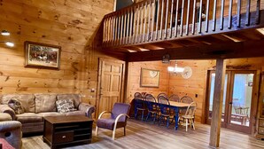 Ein gemütliches Blockhaus-Interieur mit Holzboden, einer Couch, einem Esstisch mit Stühlen und einer Treppe.