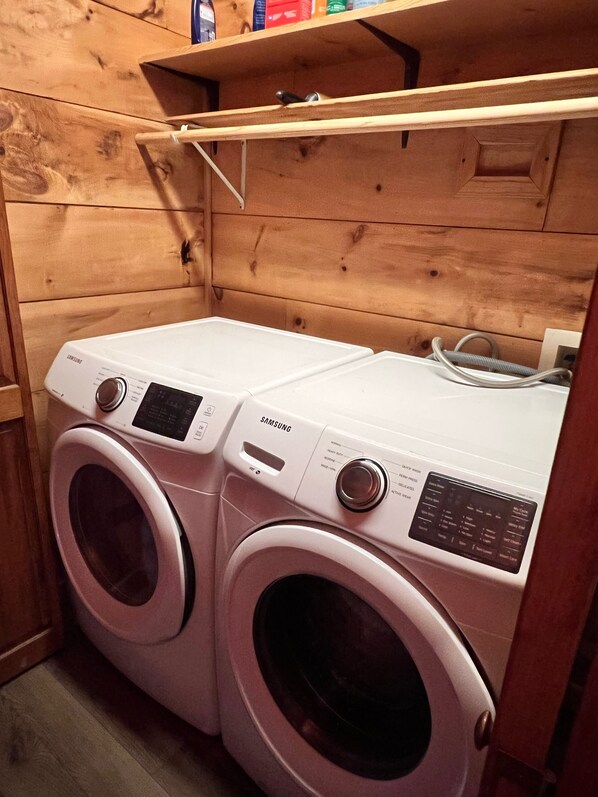 A laundry area with two white Samsung front-loading washing machines, wooden walls, and a shelf above the machines.