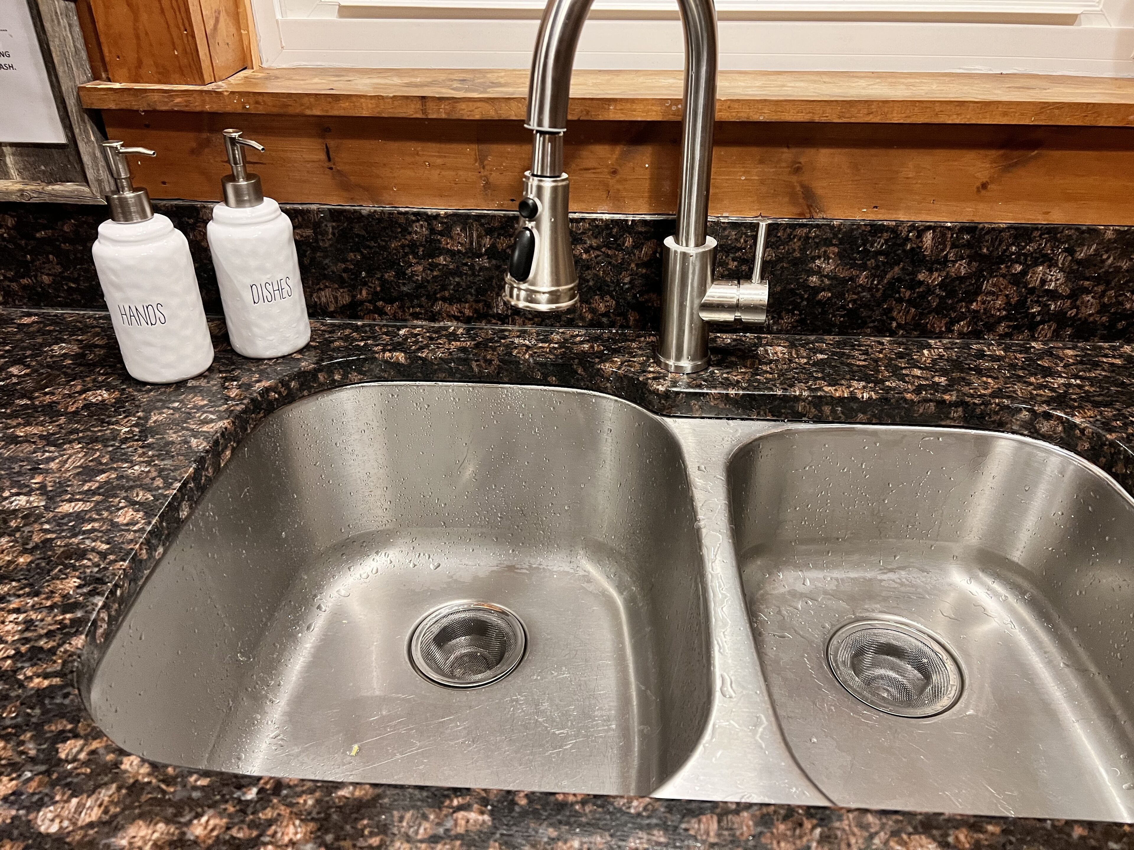 Un lavamanos de acero inoxidable con doble pileta, un grifo y dos dispensadores de jabón etiquetados como 