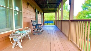 Eine hölzerne Veranda mit einem Schaukelstuhl und einer Bank, ein überdachter Bereich mit Dach und Blick auf Bäume im Hintergrund.