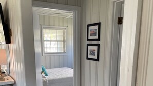 A narrow hallway leading to a bedroom with a bed, a window with blinds, and a door with a picture frame on the wall.