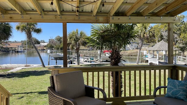 A covered outdoor deck with wooden railings, a cushioned seating area, and a view of a marina with boats and a lake.