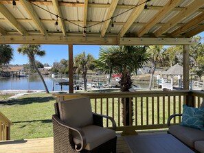 Una terraza cubierta al aire libre con barandas de madera, una zona de estar con cojines y vistas a un puerto deportivo con barcos y un lago.