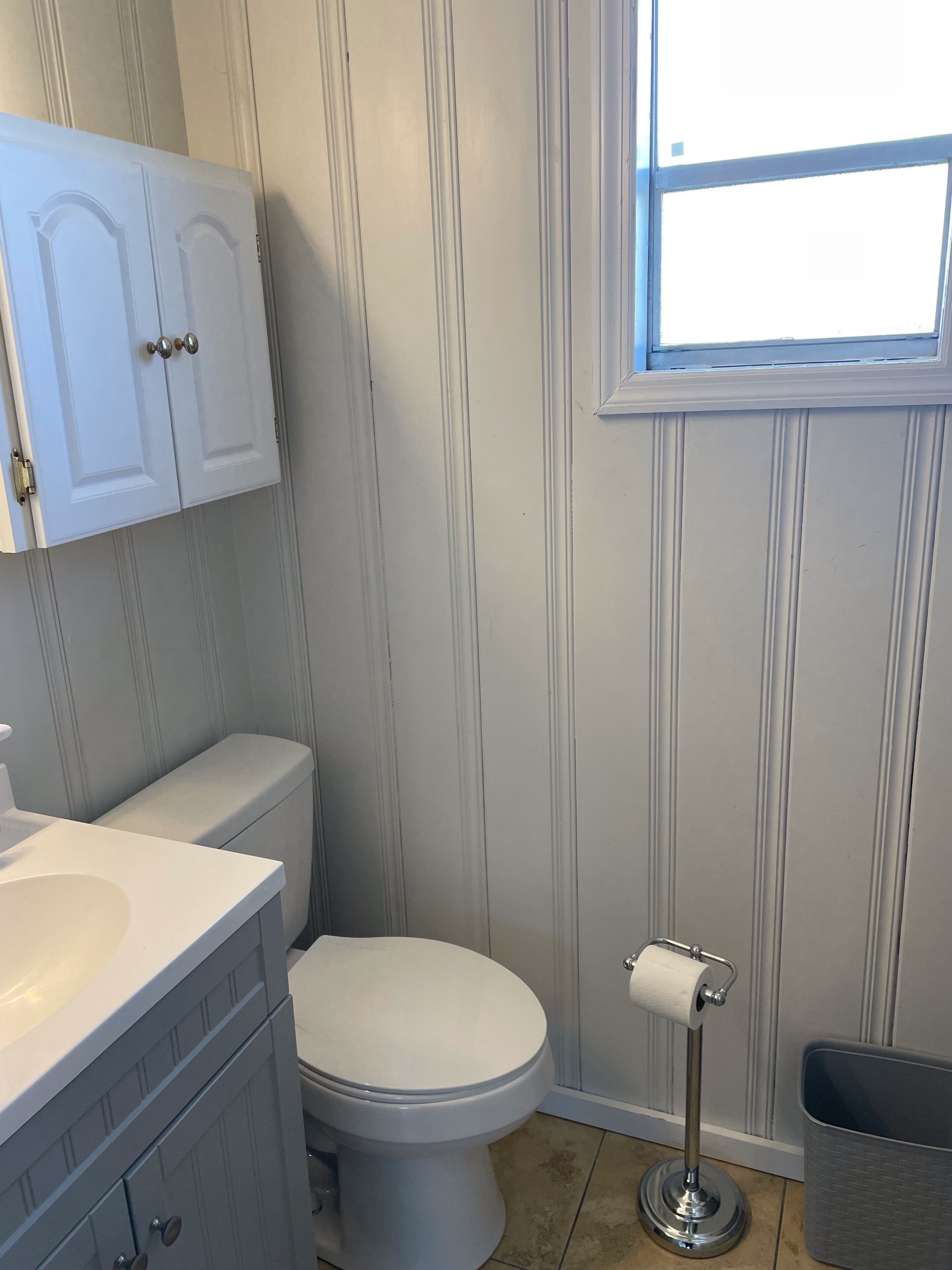 A bathroom with a toilet, a sink, a cabinet, a window, and a trash bin.