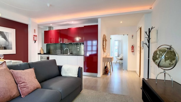 A modern living room with a grey sofa, a red kitchen in the background, and a globe on a stand.