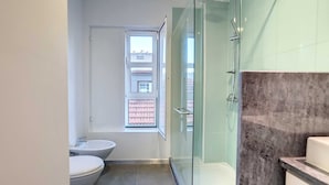 A modern bathroom with a glass shower enclosure, a toilet, and a bidet. A window provides a view of the outside.