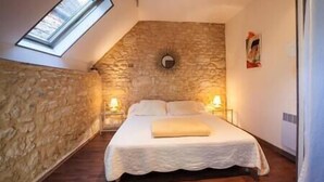 Un dormitorio con pared de piedra, tragaluz y cama con colcha blanca, además de dos lámparas de noche.