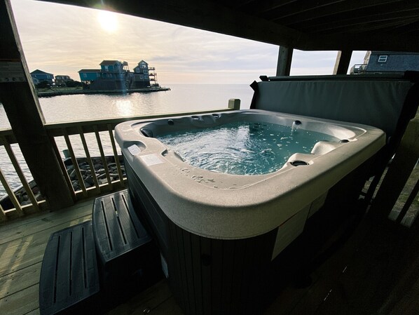 A hot tub on a wooden deck with a view of a pier and buildings across the water.