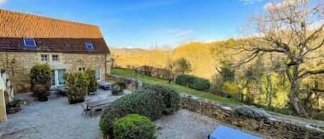 A stone building with a tiled roof, a gravel courtyard, and a view of trees and hills.
