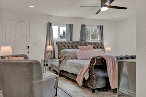 Un dormitorio con cabecera capitonada, cama con manta rosa, dos lámparas de noche, una silla y un ventilador de techo.