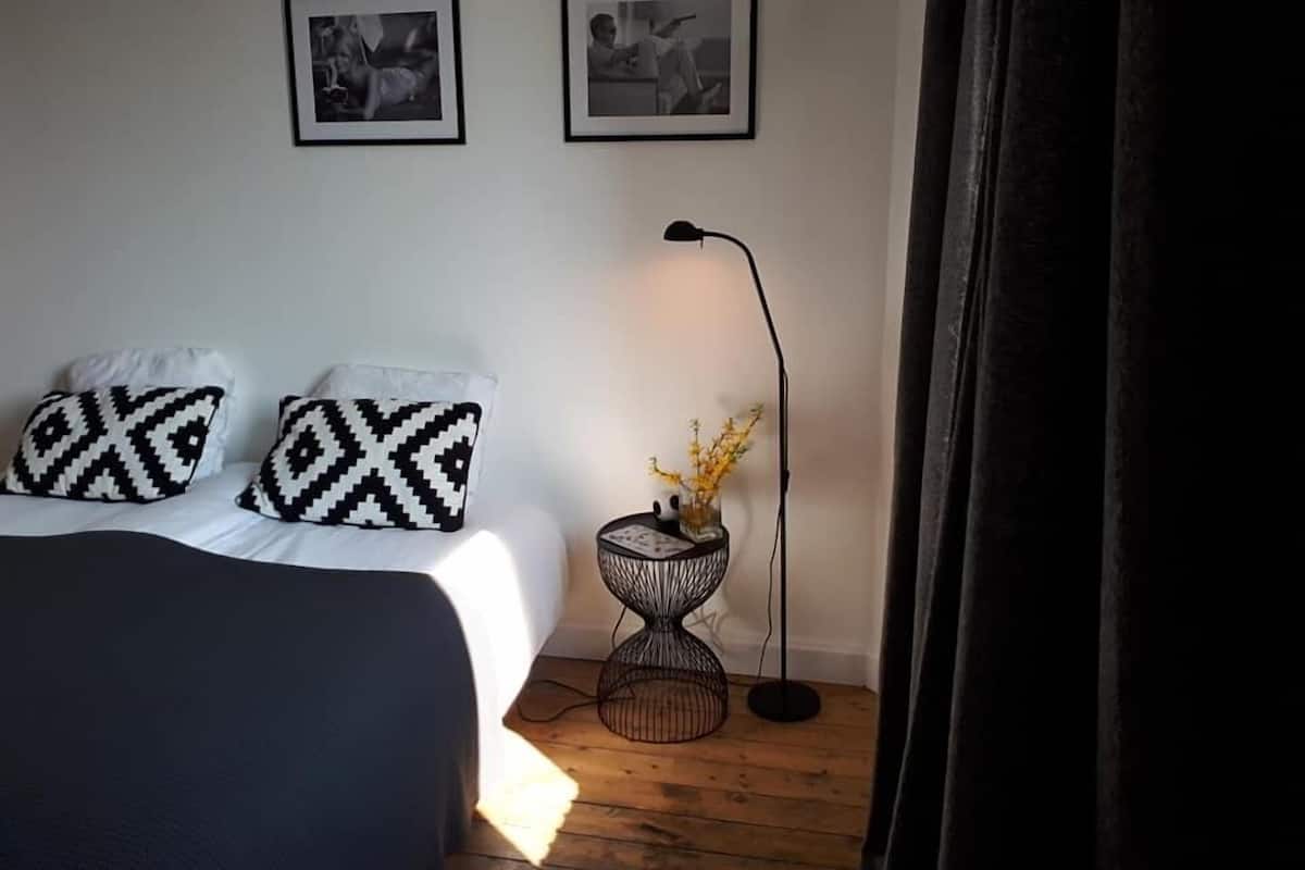 Une chambre à coucher avec un lit, deux coussins à motifs noir et blanc, une petite table ronde avec une lampe, et deux photographies encadrées accrochées au mur.