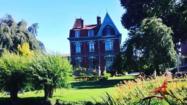 A large, red brick building with a steep roof and multiple windows, surrounded by lush greenery and a pond. - Villa Marguerite (Saint-Omer)