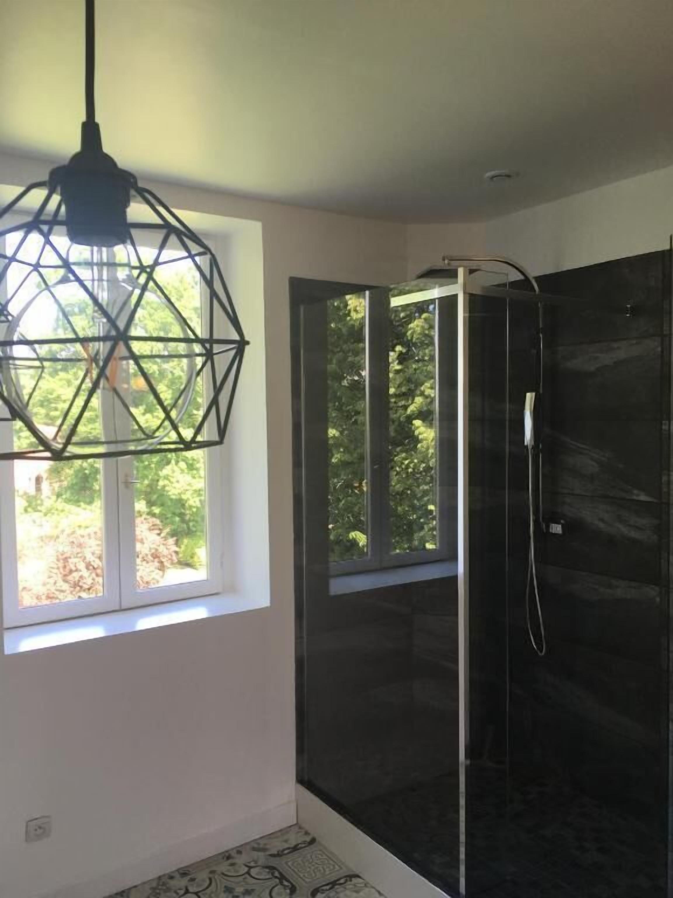 A modern bathroom with a glass shower enclosure, a geometric pendant light, and a window with a view of greenery.