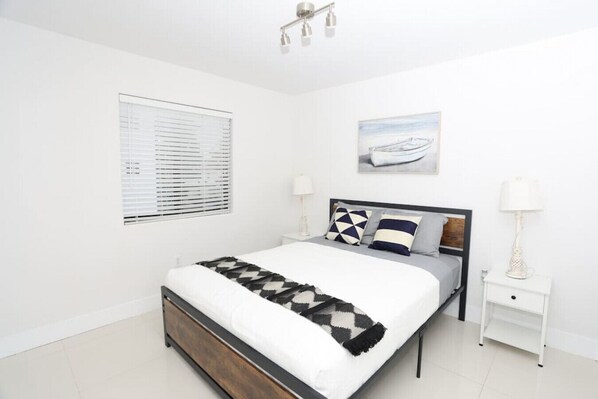 Une chambre à coucher moderne avec un grand lit, une table de chevet, une fenêtre avec des stores et un luminaire au plafond.