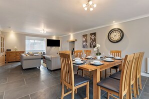 A dining area with a wooden table set for four, surrounded by wooden chairs with black cushions. A flat-screen TV is mounted on the wall above the table.