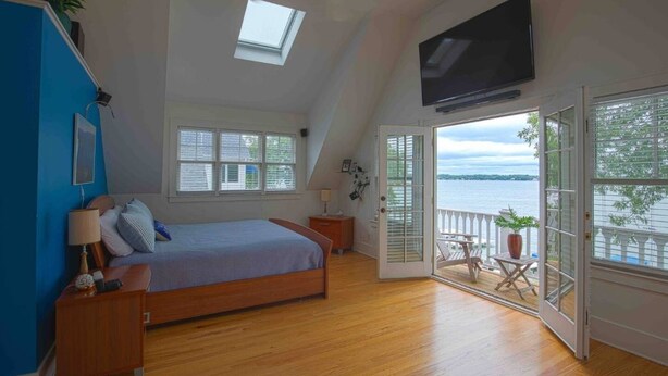 A bedroom with a large bed, a skylight, a TV, and a balcony with a view of the water. - Beautiful 3600sqft Cottage on Lake Maxinkuckee in Culver Indiana. (Culver)