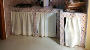 A compact kitchen with a gas stove, a white microwave, and a sink. A shelf above the counter holds various kitchenware and a few glasses. The backsplash features a geometric tile pattern.
