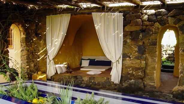 A stone-walled room with a bed, white curtains, and a small table with a bottle.